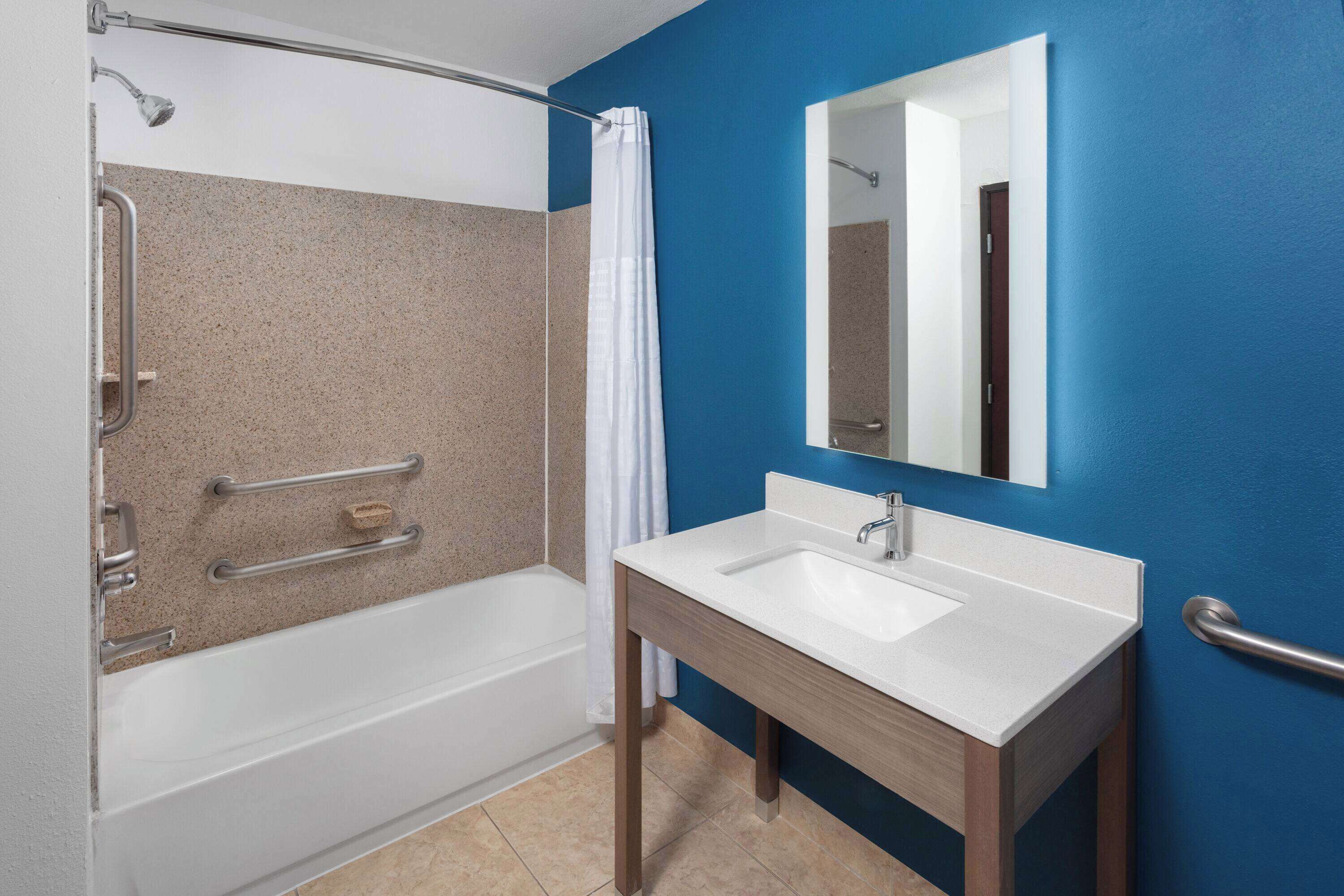 Un baño con bañera, ducha con barras de apoyo, lavamanos con espejo y pared azul.