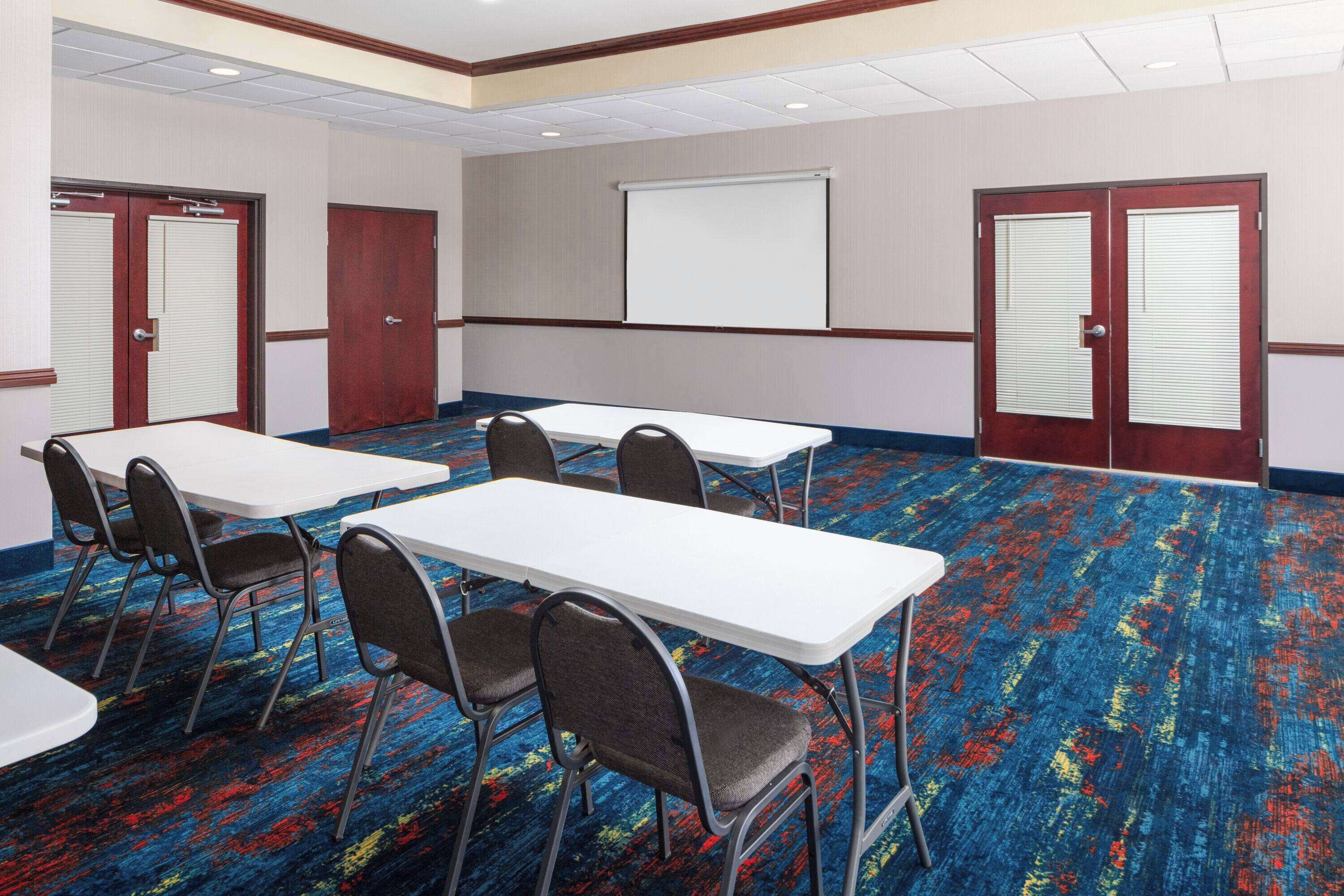 Une salle de conférence équipée de tables et de chaises, d’un écran de projection et de portes.