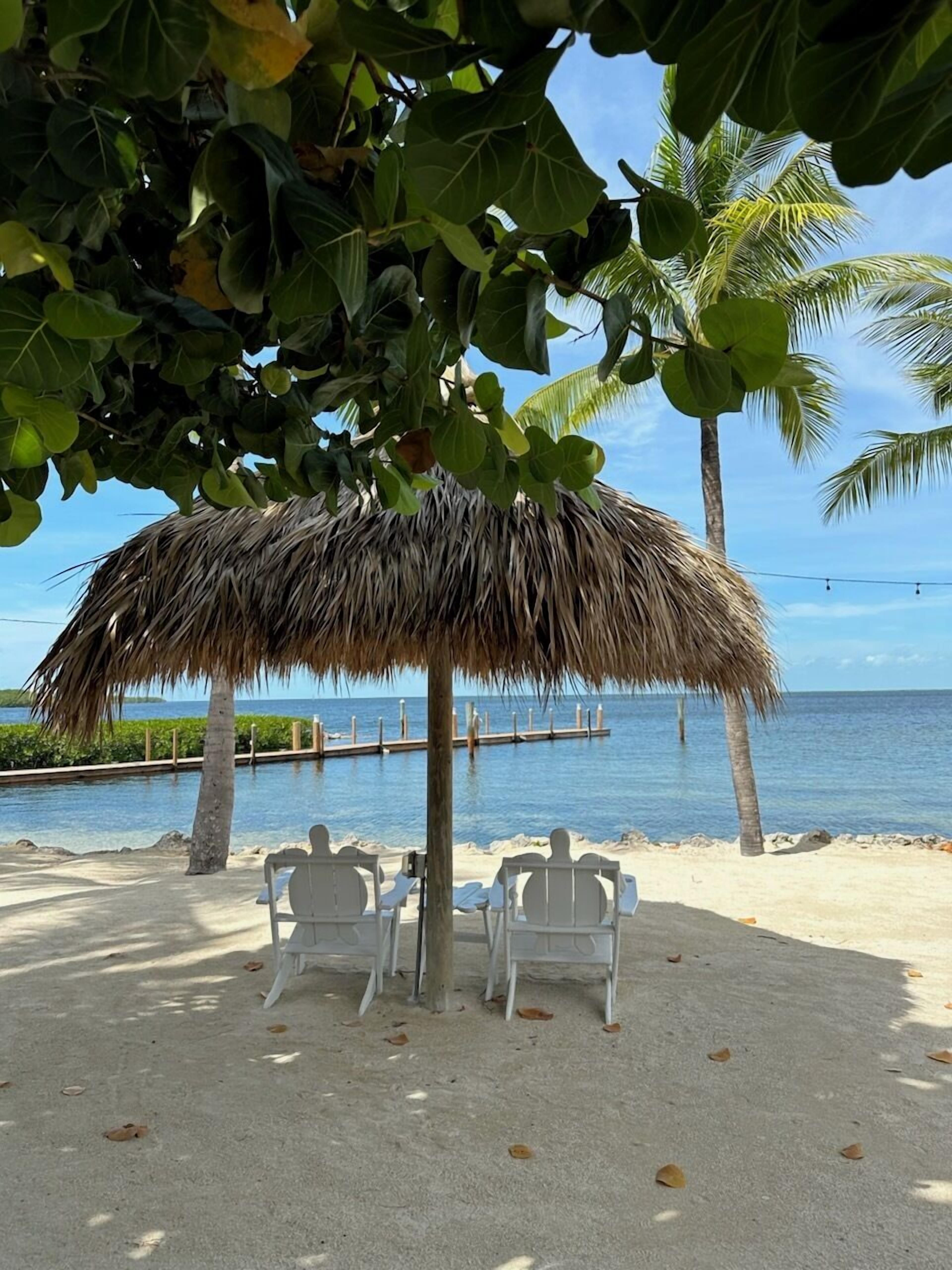A beach with a thatched umbrella, white chairs, and a view of the ocean.