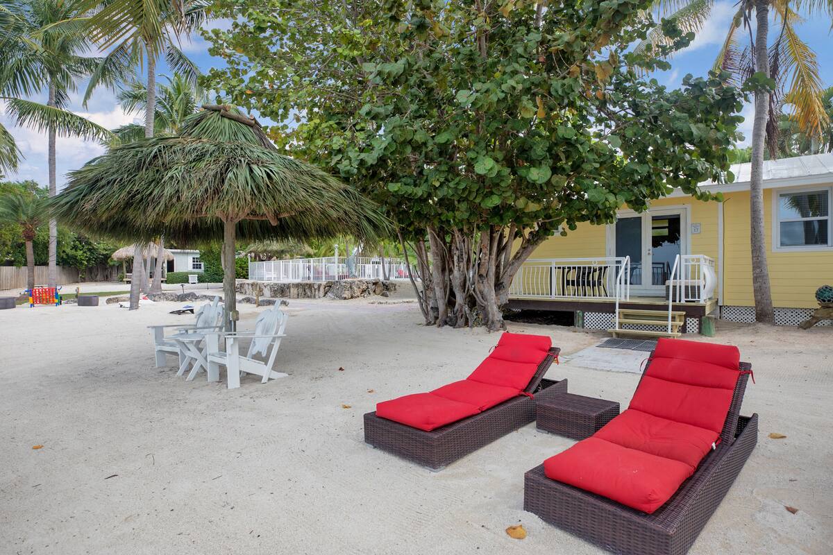 A beachside lounge area with wicker lounge chairs and a thatched umbrella, a small pool, and a building with a porch.