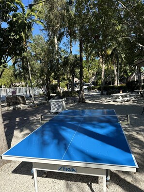An outdoor ping pong table with a blue playing surface and a white frame, surrounded by trees and a gravel area. - Atlantic Bay Resort (Tavernier)