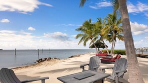 Un espace de détente aménagé sur la plage, avec des fauteuils Adirondack et une table, des palmiers et une vue sur l’océan.