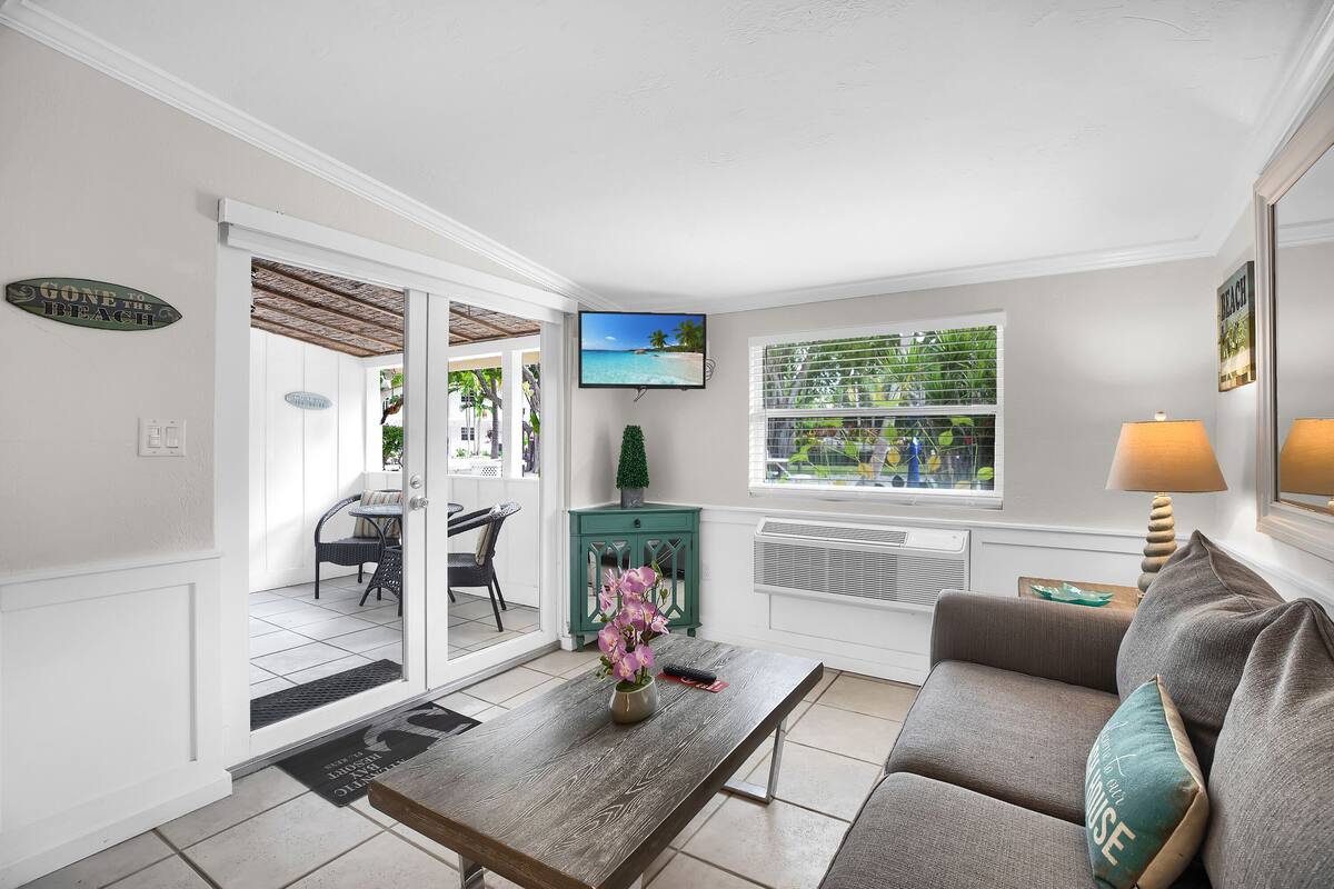A living room with a sofa, coffee table, and a TV. There's a window with blinds and a small plant on a cabinet.
