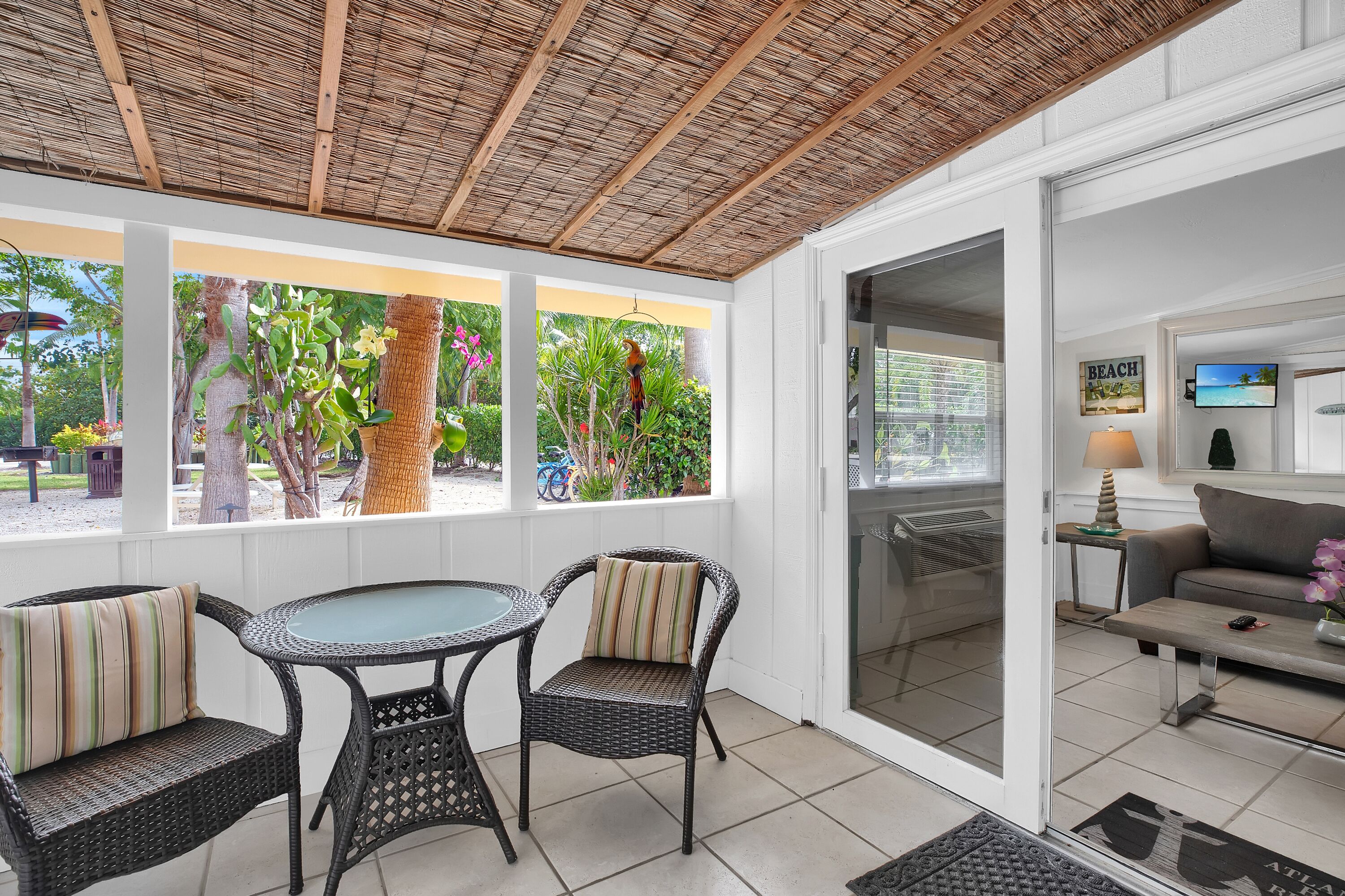 A covered patio with wicker furniture, a glass table, and a view of tropical plants and a beach scene.