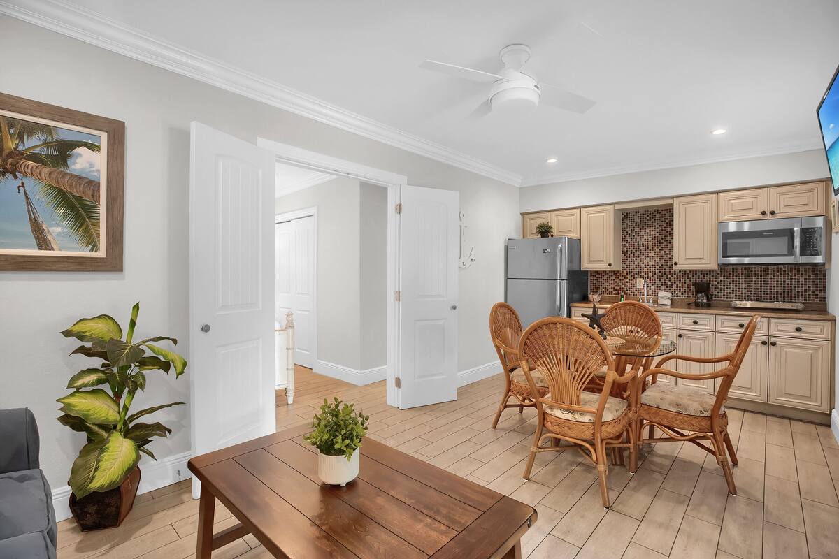 A modern kitchen with white cabinets, a microwave, and a refrigerator. There is a dining area with a wooden table and wicker chairs.