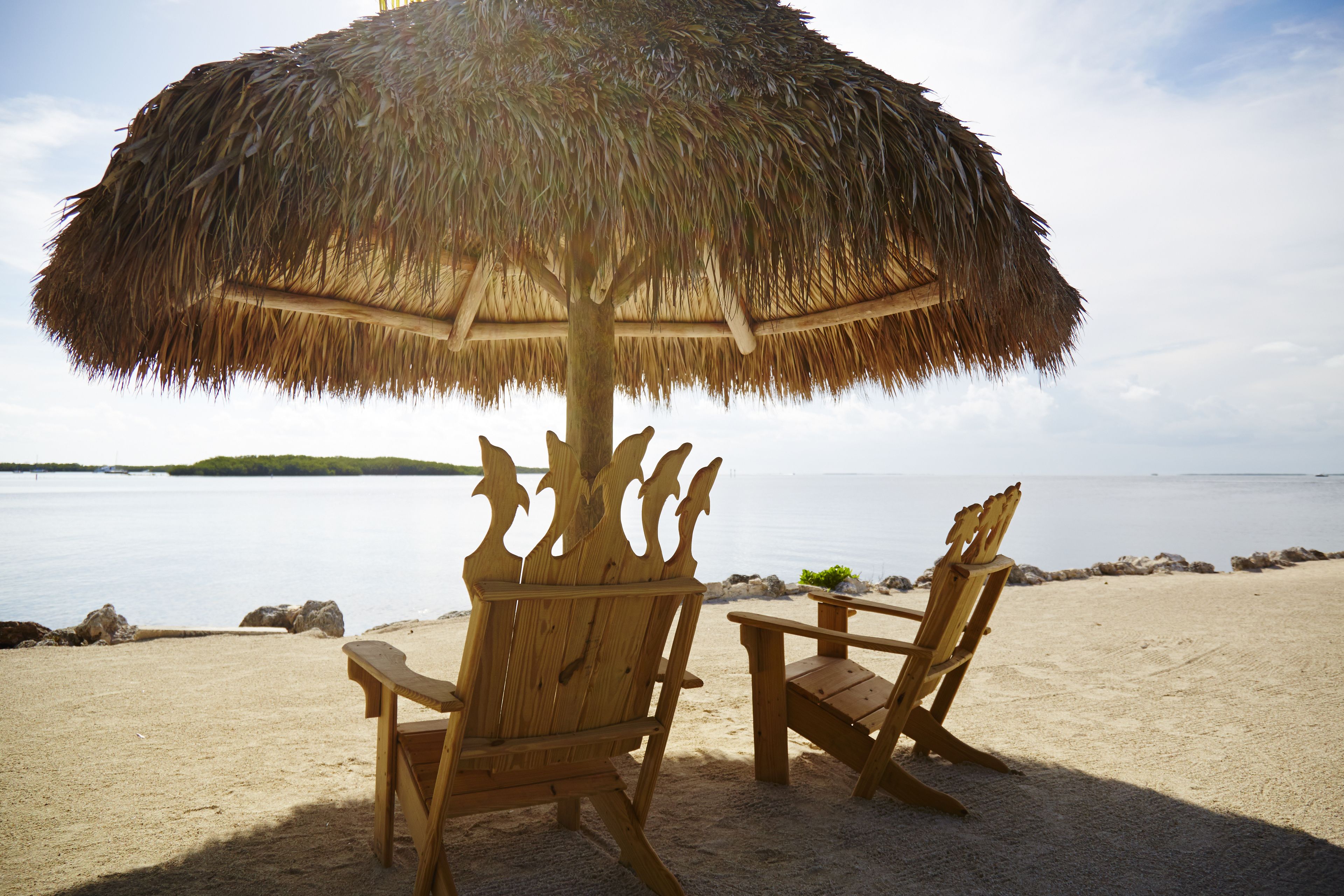 A beach scene with a thatched umbrella and two wooden chairs.