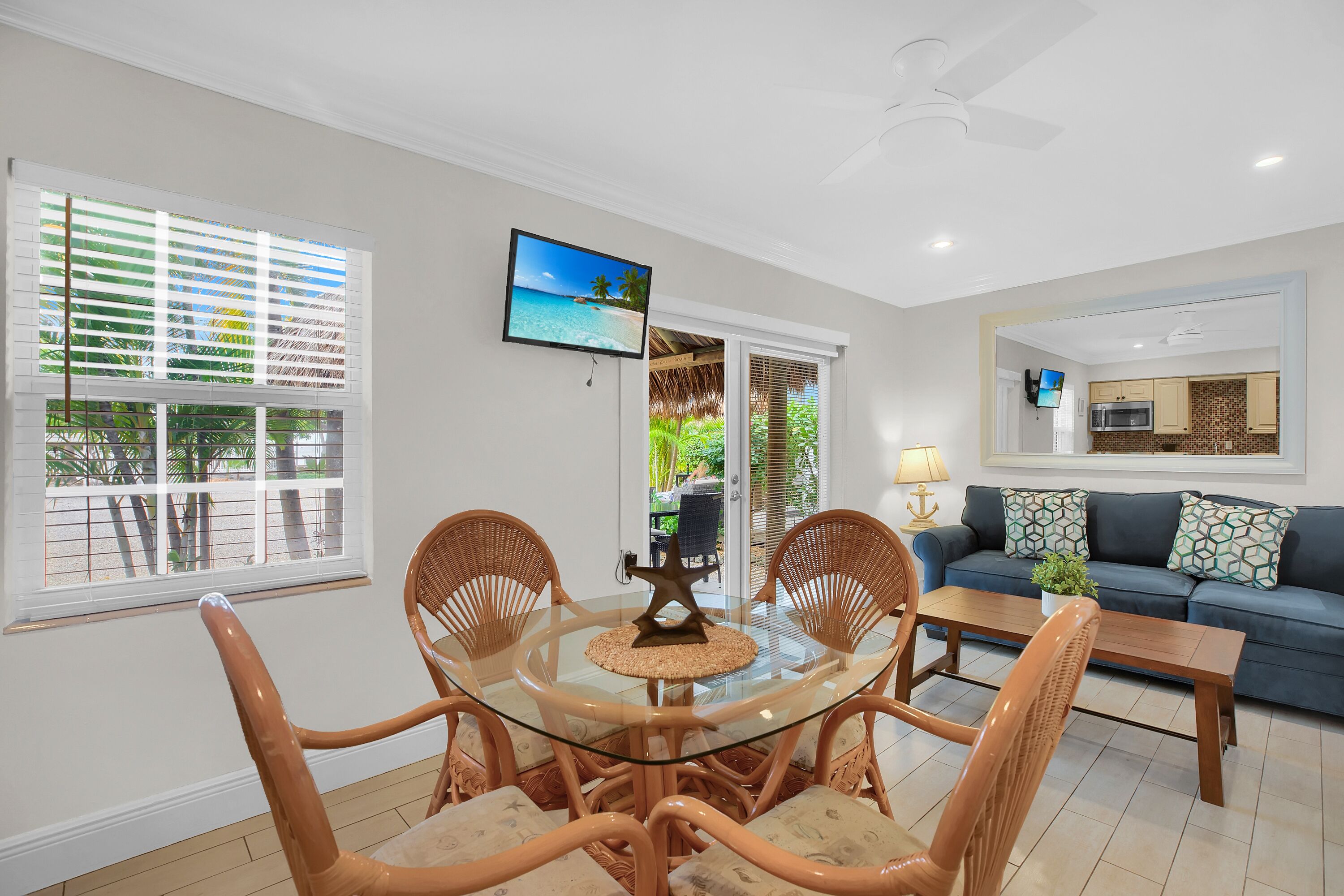 A dining area with a glass table and wicker chairs, a living room with a sofa and TV, and a kitchen with a microwave.