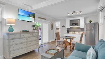 Ein großzügiger Wohnraum mit einem blauen Sofa, einem Couchtisch und einem Fernseher, der eine Strandansicht zeigt. Im Hintergrund ist eine Küche mit weißen Schränken und einem Edelstahl-Kühlschrank erkennbar.