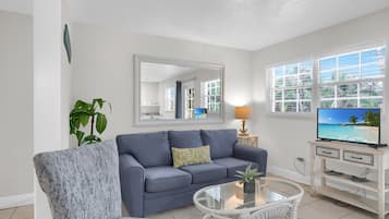Una sala de estar con un sofá gris, una mesa de centro redonda de vidrio, un televisor sobre un mueble blanco y un ventanal con vista a palmeras.