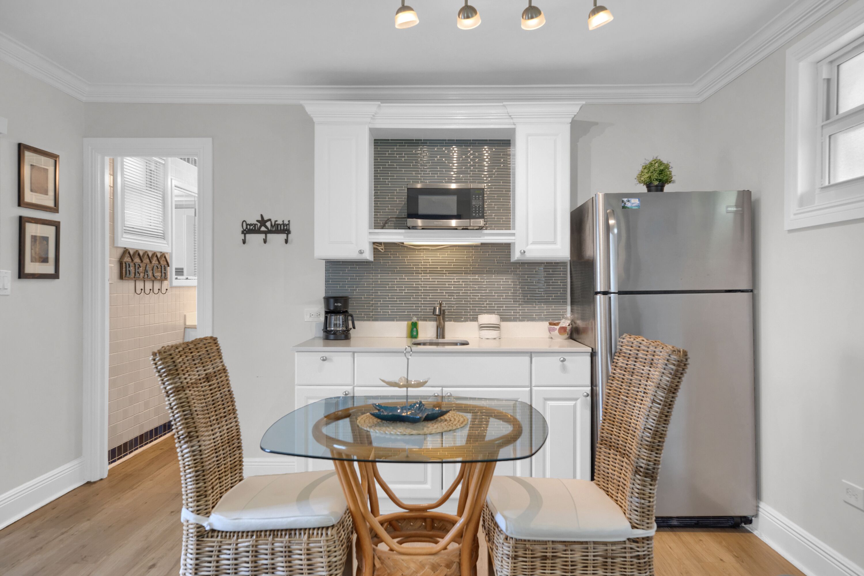 A modern kitchen with white cabinetry, a glass-top dining table, wicker chairs, a stainless steel refrigerator, and a microwave above the stove.