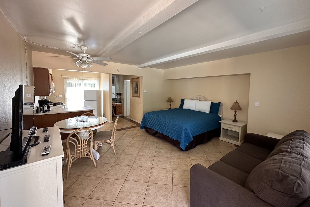 Une chambre d’hôtel compacte avec une kitchenette, un lit avec une literie bleue, un canapé marron et un ventilateur de plafond.