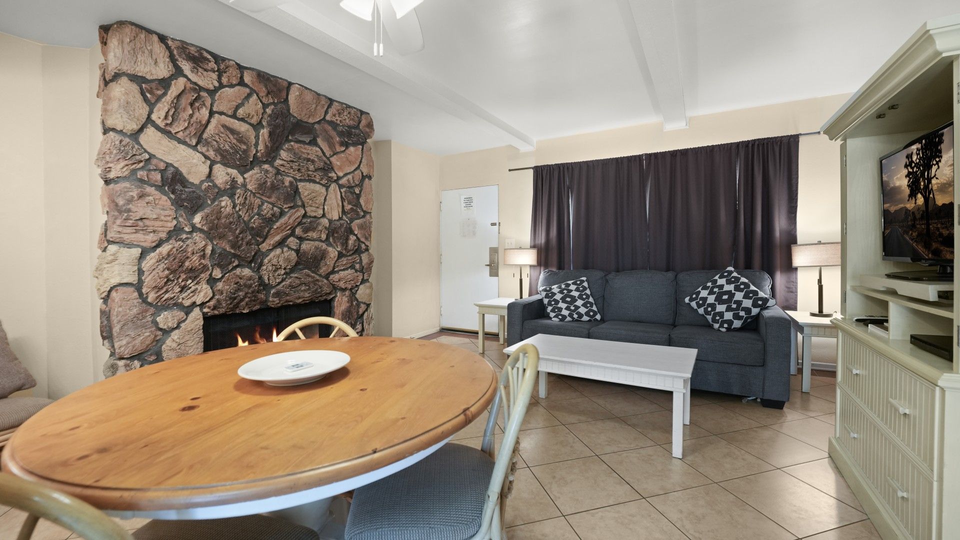 Uma sala de estar com lareira de pedra, mesa de jantar, sofá com almofadas estampadas e televisão sobre um aparador.