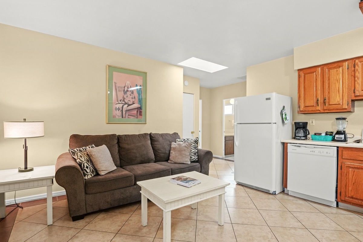 A compact living space with a sofa, a coffee table, a lamp, a refrigerator, a dishwasher, and wooden cabinets.