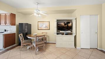 A compact kitchen with a refrigerator, a dining table with chairs, a ceiling fan, and a television on a cabinet.