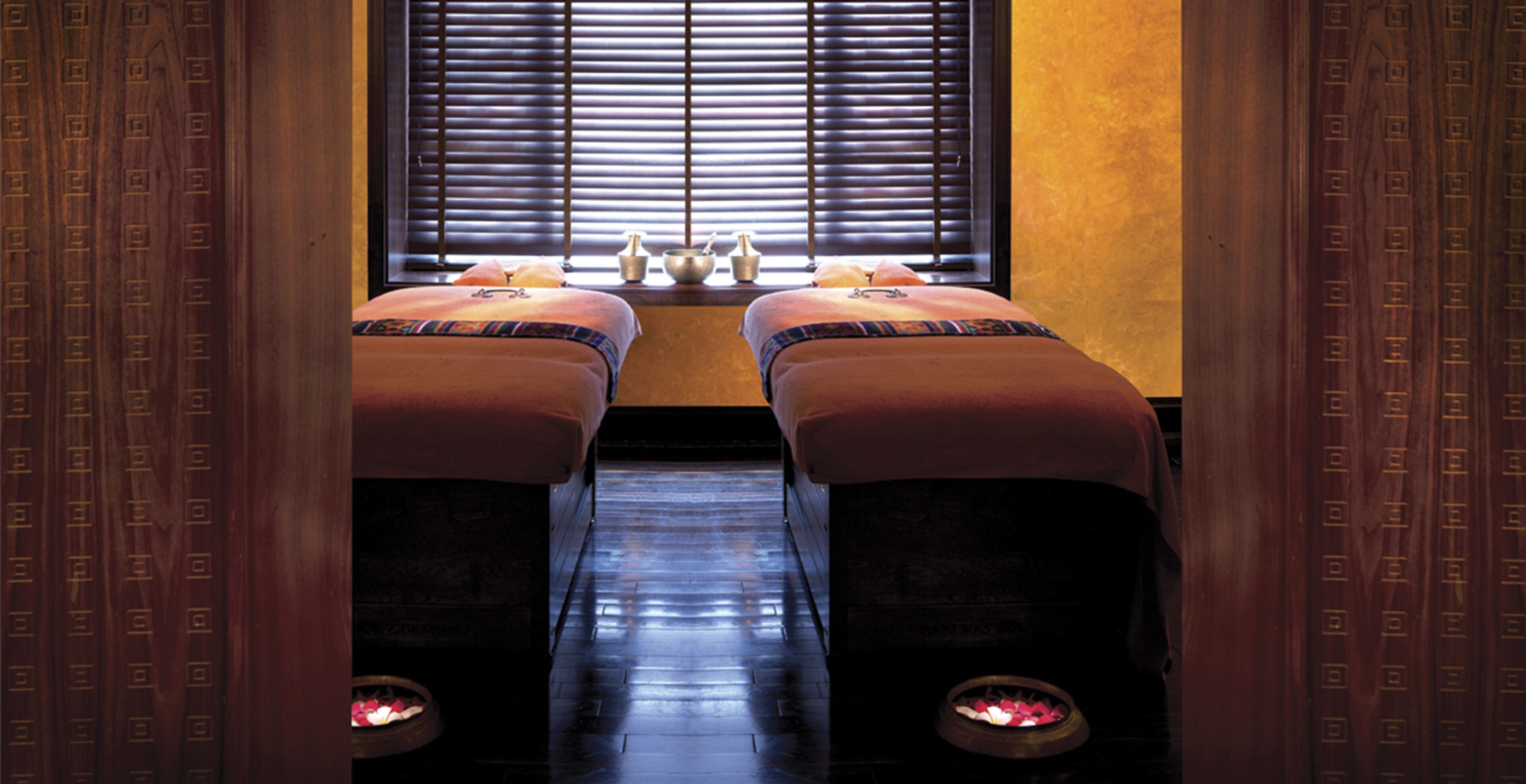 Une pièce comprenant deux tables de massage, une fenêtre avec des stores et une petite table ornée de bougies.