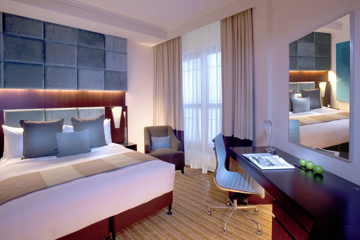 A hotel room with a large bed, a desk, a chair, a mirror, and a window with curtains.