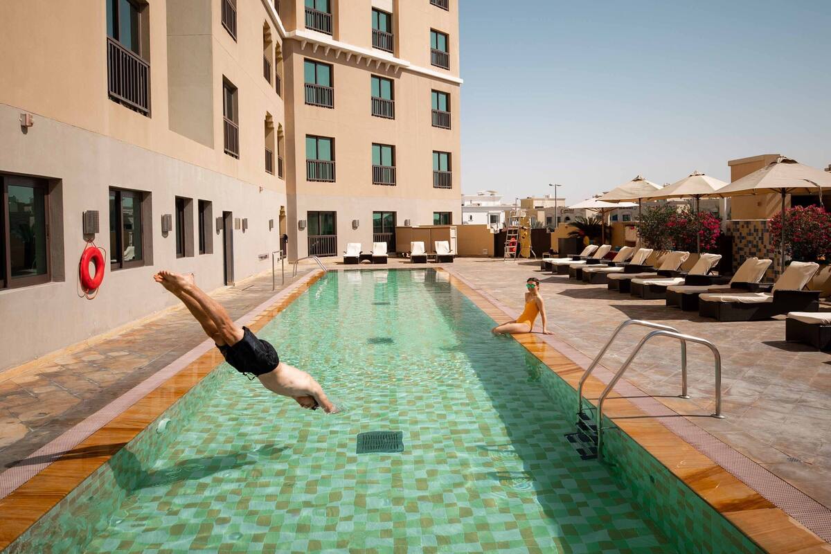 A swimming pool with a diving board, lounge chairs, and a high-rise building in the background.