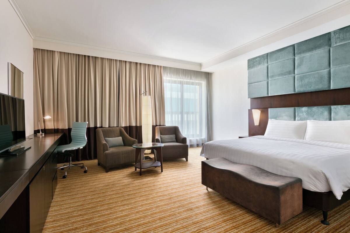 A hotel room with a large bed, two armchairs, a desk with a computer, a lamp, and a large window with curtains.