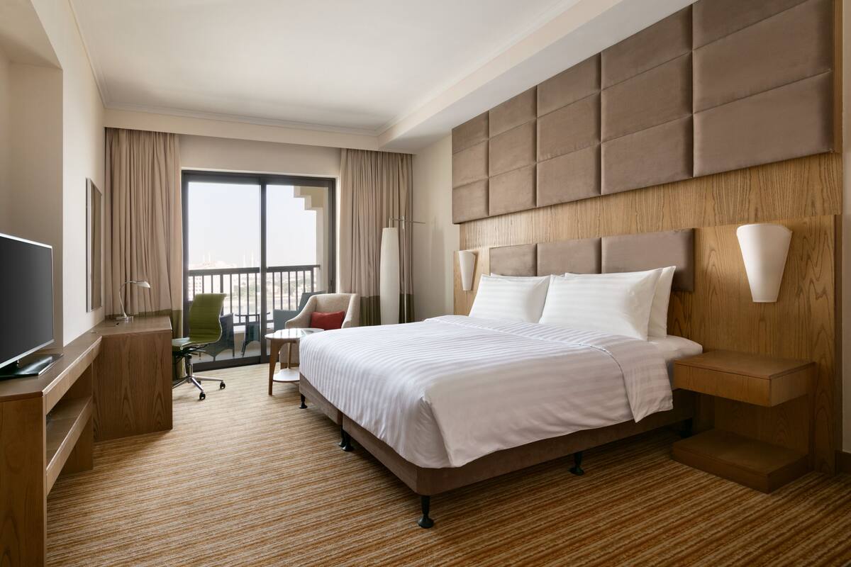 A hotel room with a large bed, a desk with a TV, a chair, a lamp, and a view of the outdoors.
