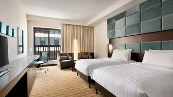 Quarto de hotel com duas camas, uma mesa, uma cadeira, uma TV e uma janela grande com cortinas.