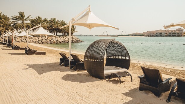 A beach with sun loungers and a large wicker chair, a white umbrella, and a rocky shoreline.
