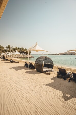 A beach with sun loungers and a large wicker chair, a white umbrella, and a rocky shoreline. - Traders Hotel, Qaryat Al Beri (Abu Dhabi)