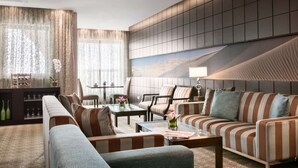A modern hotel room with a sectional sofa, armchairs, a coffee table, and a dining area with a table and chairs.