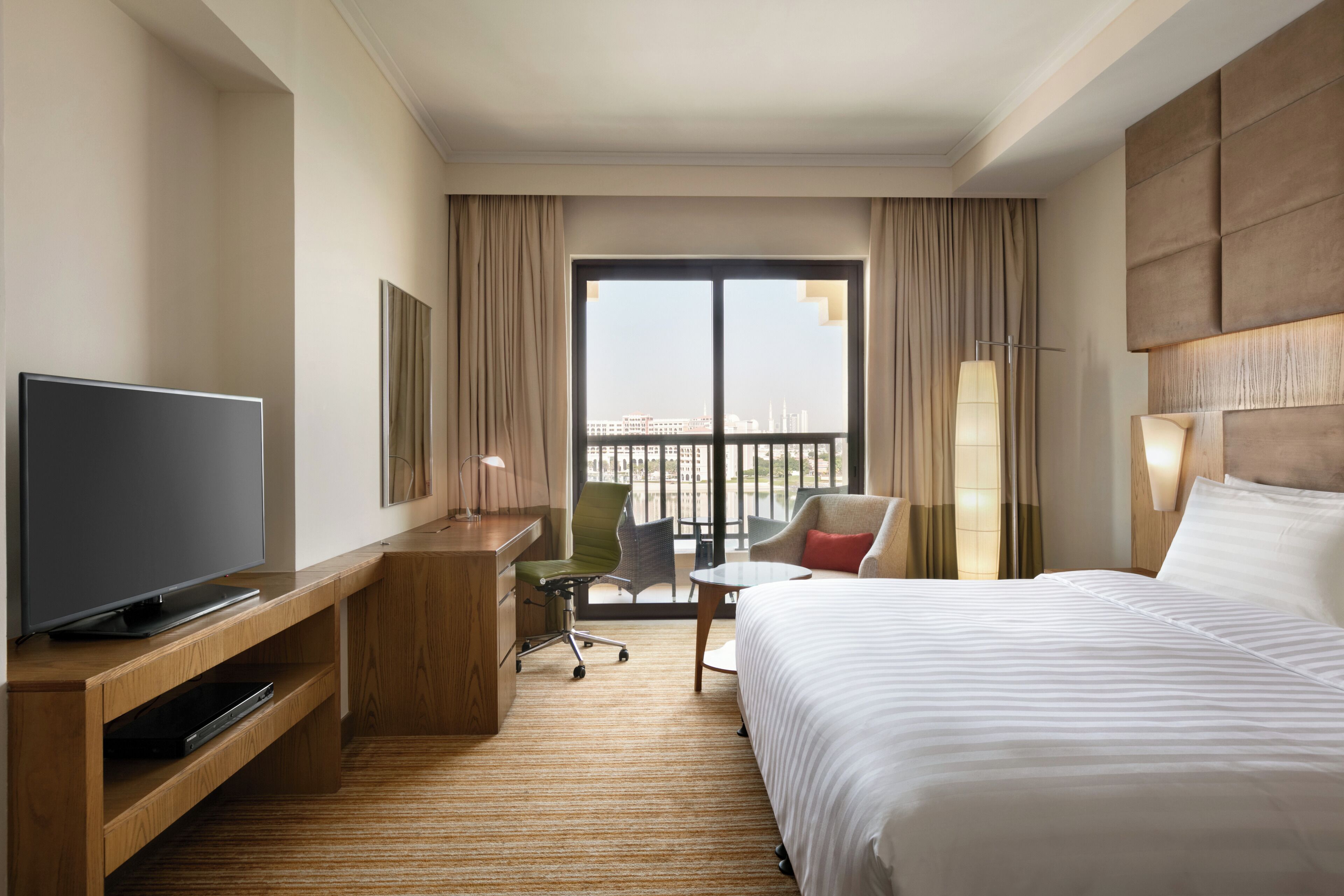 Ein Hotelzimmer mit einem Bett, einem Fernseher auf einem Holzboden, einem Schreibtisch, einem Stuhl und Blick auf die Stadt durch große Fenster.