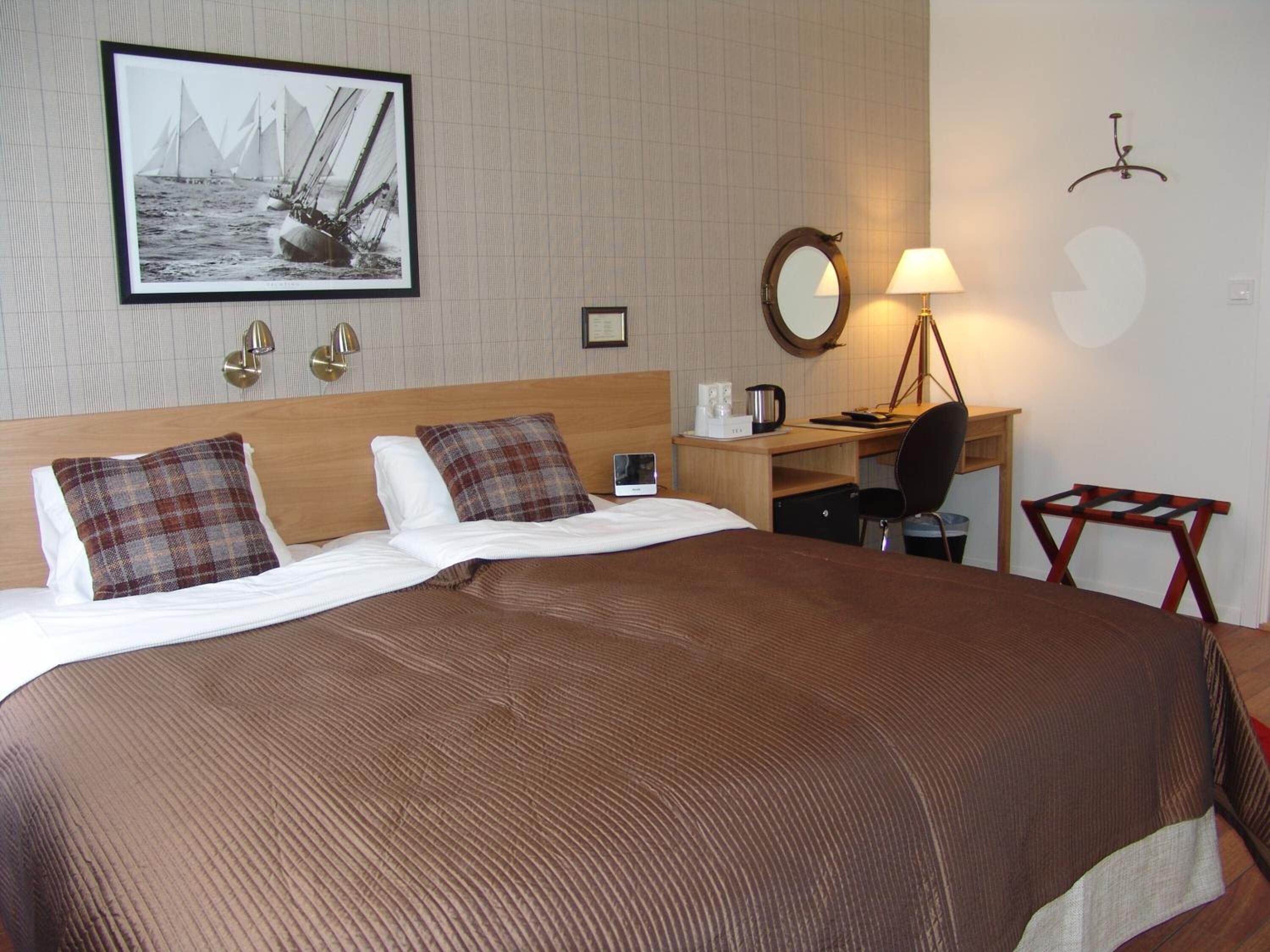 A hotel room with a large bed, a desk with a chair, a lamp, and a framed picture of sailboats on the wall.