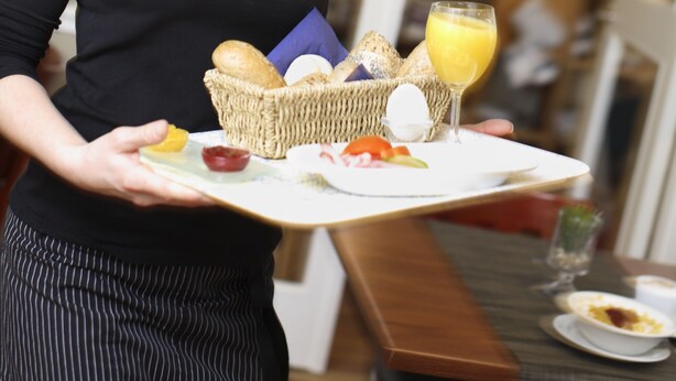 A person holding a tray with a basket of bread, a glass of orange juice, and various breakfast items.