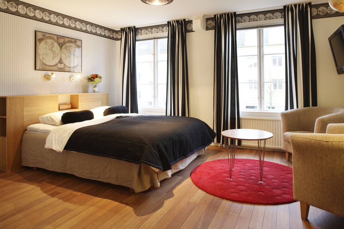 A hotel room with a bed, a small round table, a red rug, and a chair.