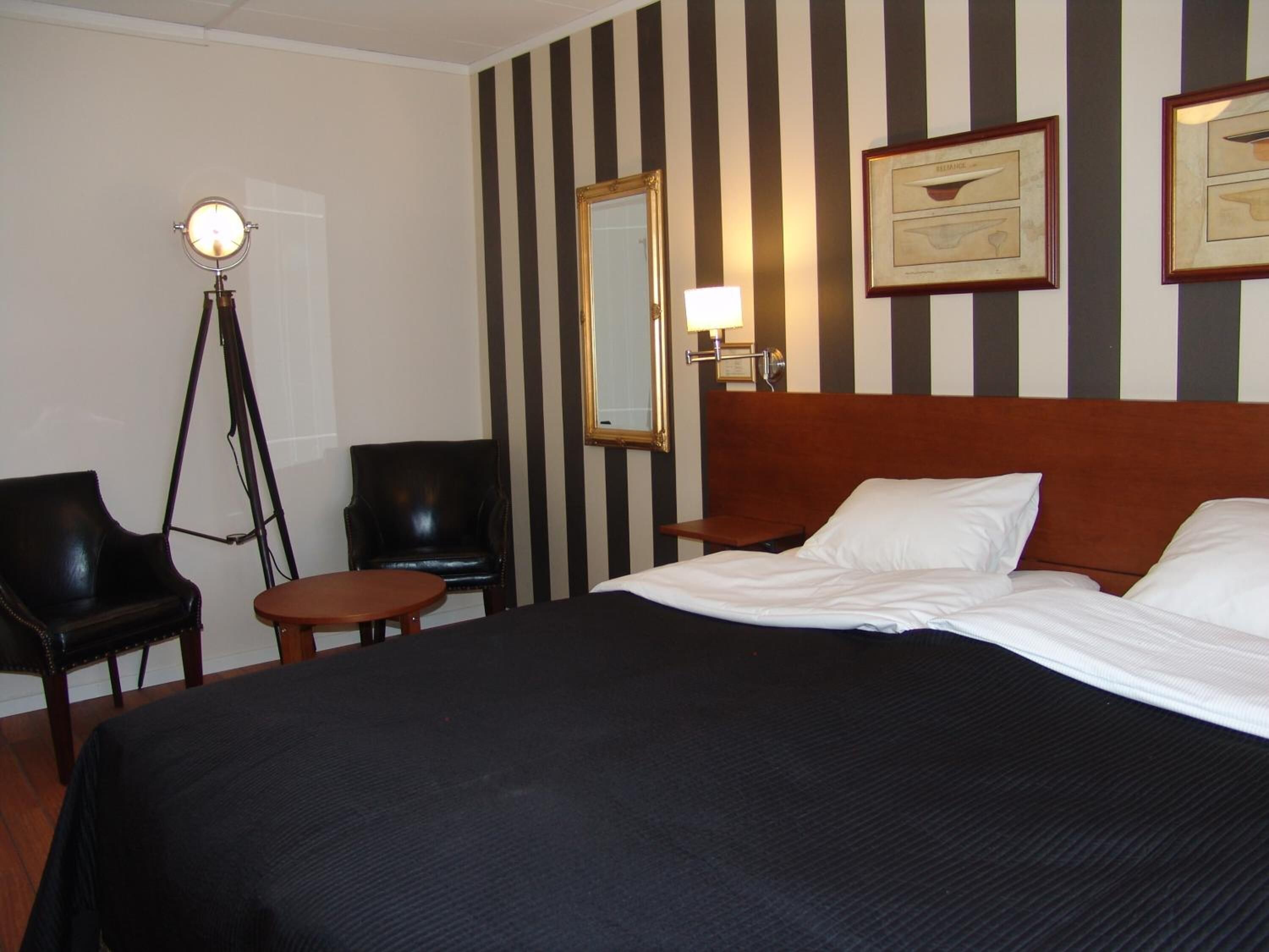 A hotel room with a large bed, two chairs, a small table, a floor lamp, and striped wallpaper.