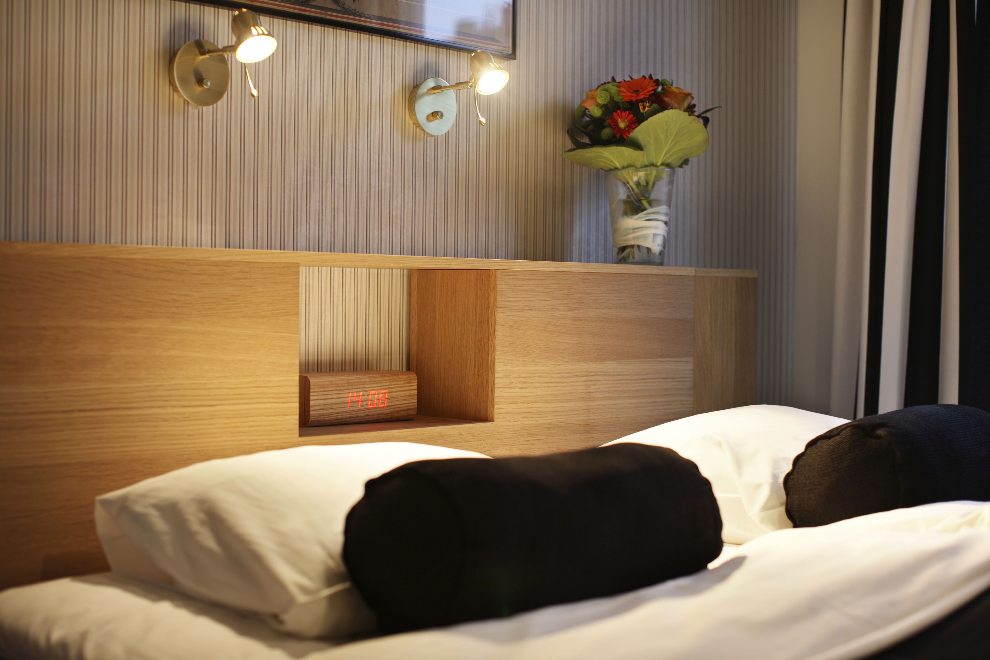 Una habitación de hotel con cabecera de madera, una mesita de noche con un reloj y un florero sobre la mesita.