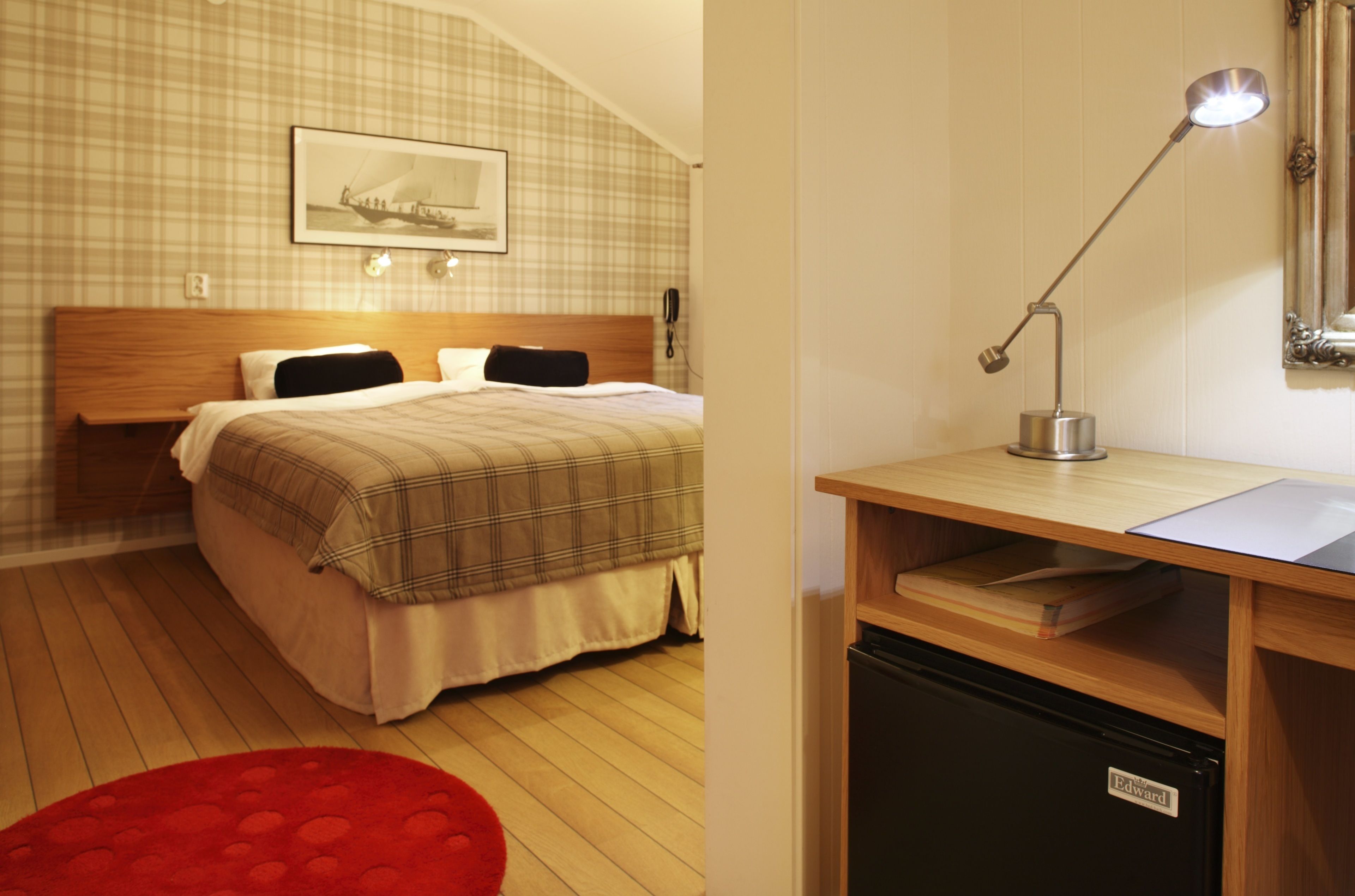 Una habitación de hotel con una cama, un escritorio con lámpara, un refrigerador pequeño y una alfombra roja.