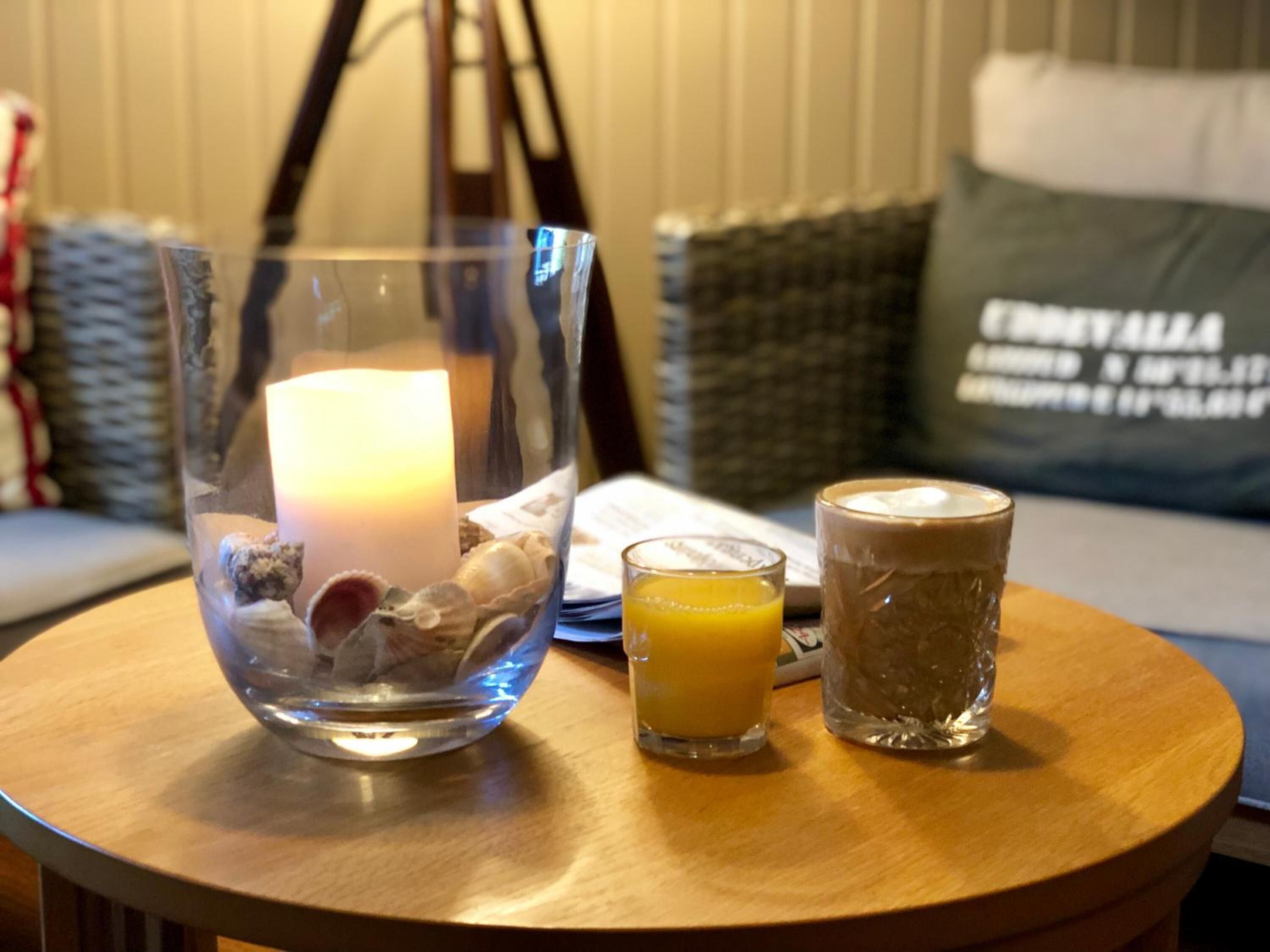 Un cuenco de vidrio transparente con una vela, dos vasos de jugo de naranja y otra vela en una habitación con una mesa de madera y un sofá al fondo.