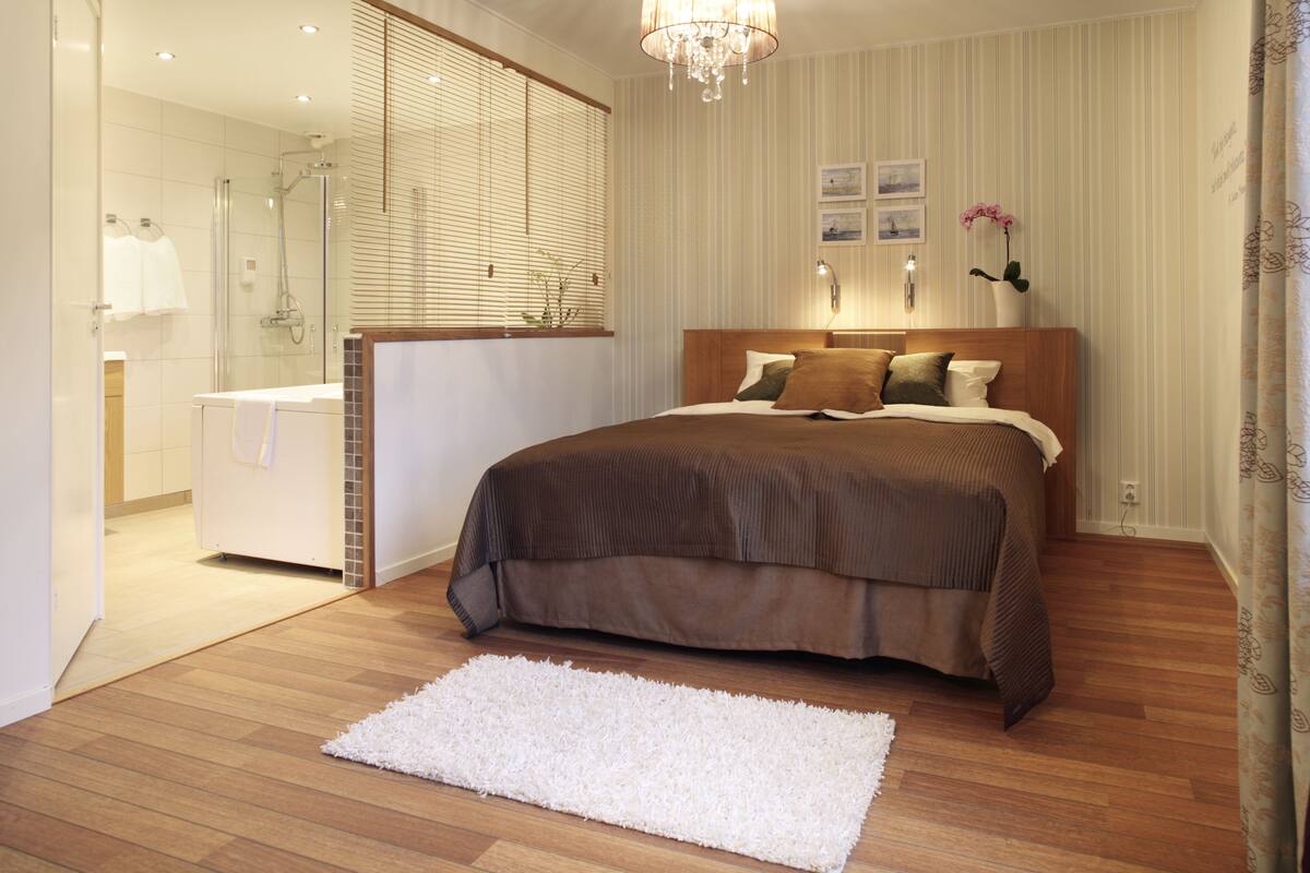 A modern bedroom with a wooden floor, a large bed with a brown headboard, a white rug, and a bathroom with a glass shower enclosure.