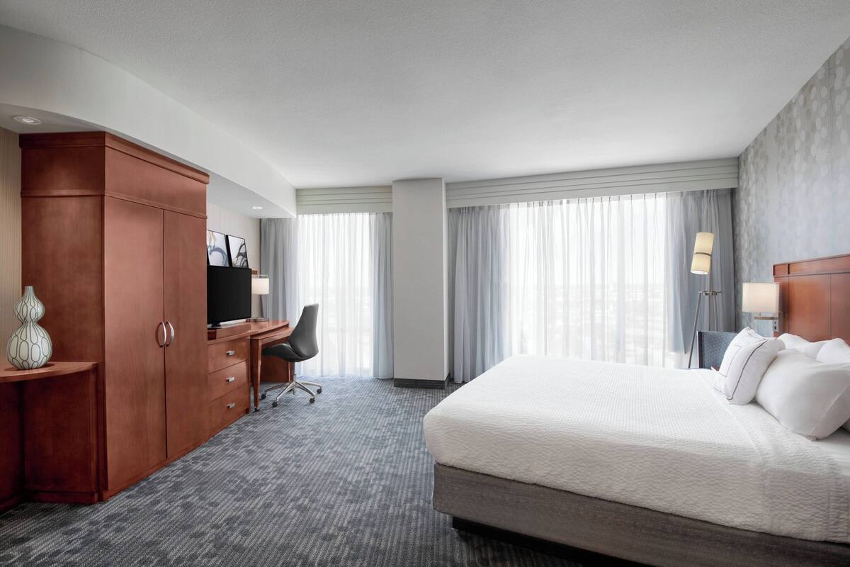 A hotel room with a large bed, a desk with a chair, a wardrobe, and a large window with curtains.
