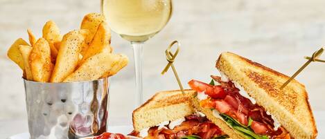 Un sandwich au bacon, à la tomate et à la laitue, un verre de vin blanc et une portion de frites servie dans un récipient en métal.