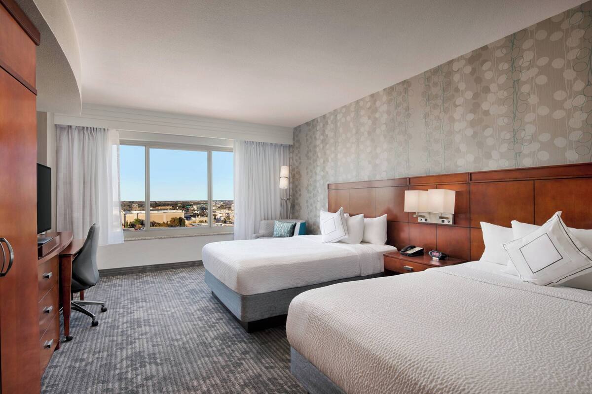A hotel room with two beds, a desk, a TV, and a large window with a city view.