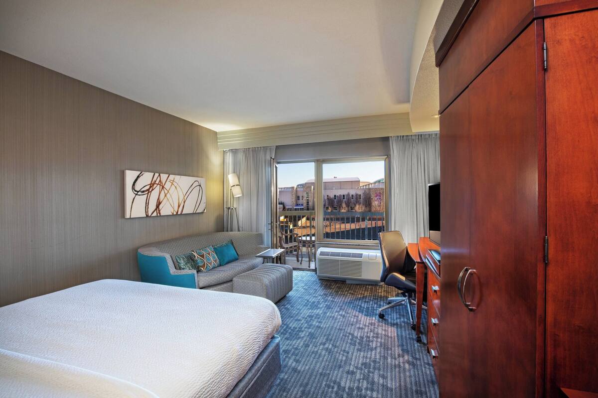 A hotel room with a large bed, a sofa, a desk, and a wardrobe. There is a window with a view of buildings and a cityscape.