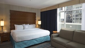 Ein Hotelzimmer mit einem großen Bett, zwei Nachttischen mit Lampen, einem Sofa, einem Fenster mit Blick auf Gebäude und einem gemusterten Teppich.