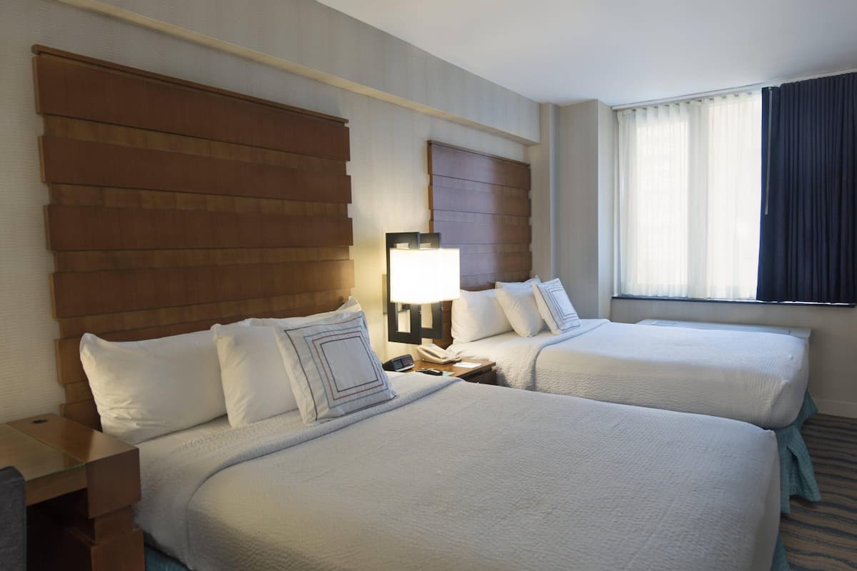 Habitación de hotel con dos camas, una mesita de noche con lámpara y una ventana con cortinas.