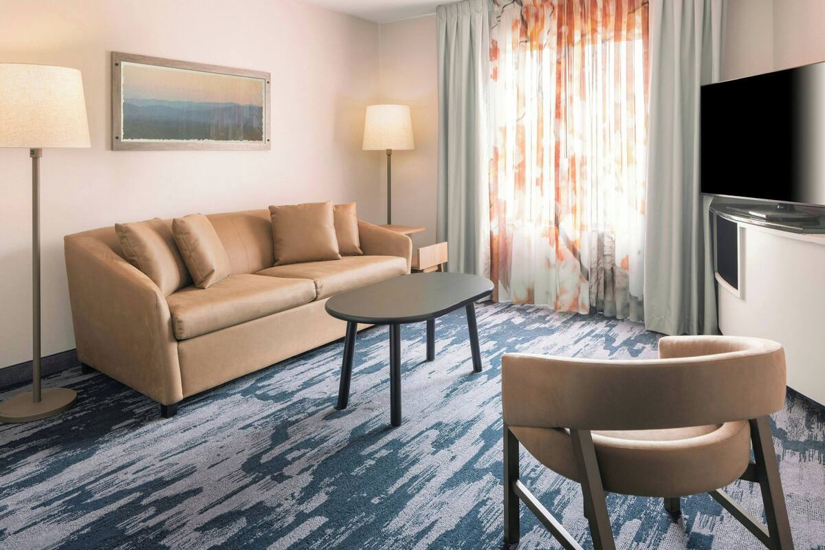 Habitación de hotel con sofá color beige, una mesita, una silla, televisión y un cuadro en la pared.
