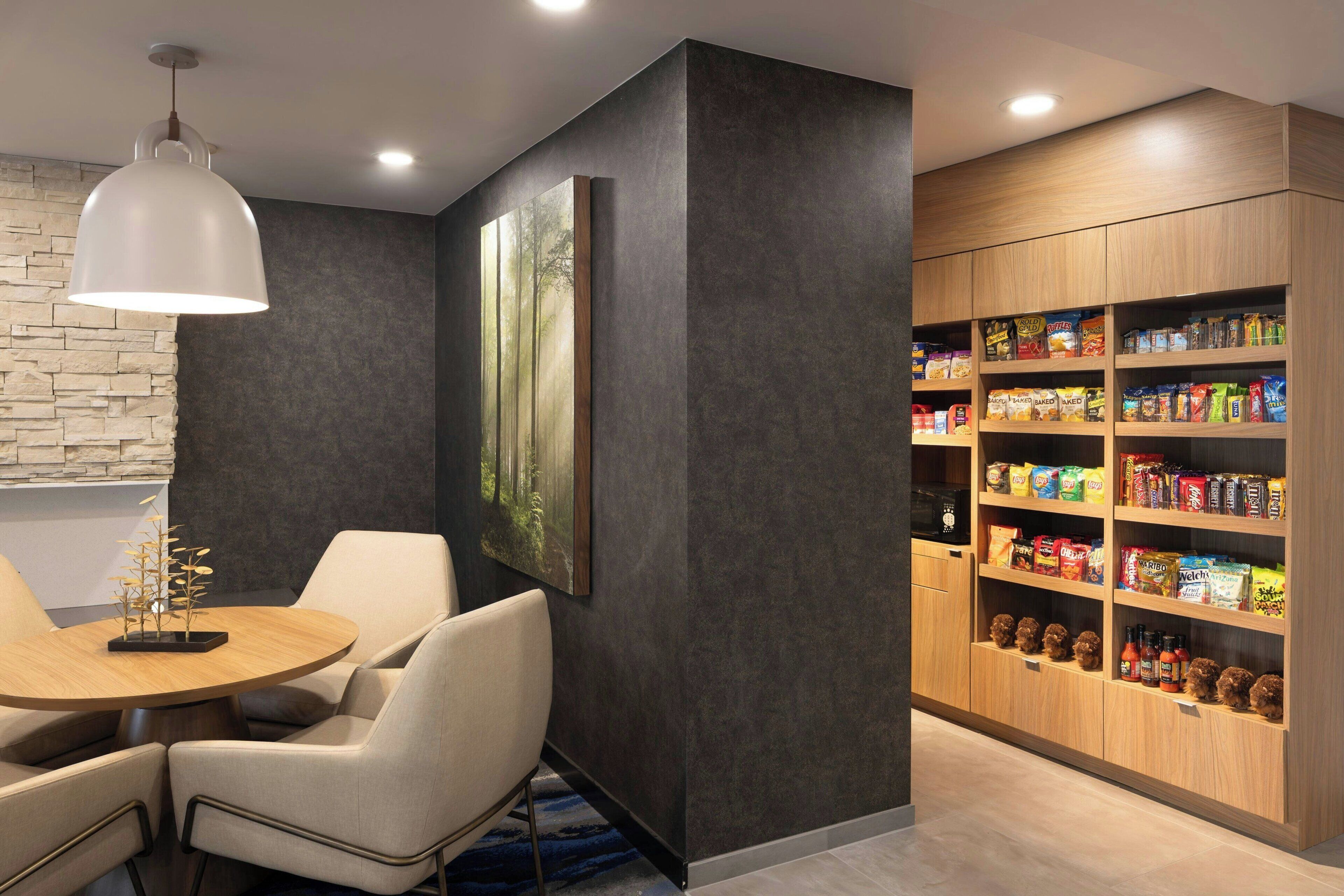 A modern dining area with a round table, chairs, and a pendant light. A large painting of a forest is on the wall, and a wooden cabinet with snacks is to the right.