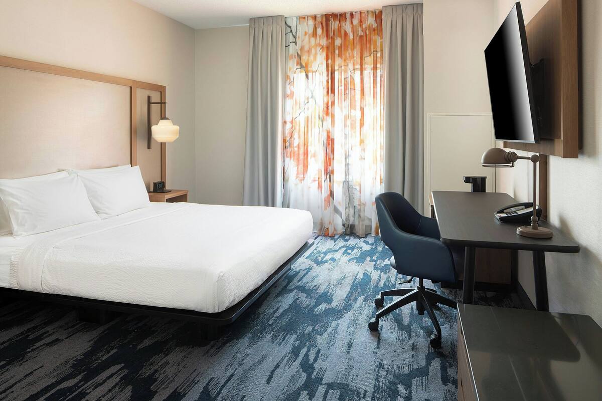 Habitación de hotel con cama, escritorio, silla, televisor y ventana con cortinas.