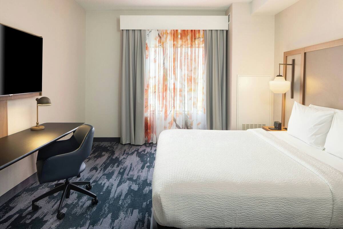 Habitación de hotel con cama, escritorio, silla, televisión y ventana con cortinas.