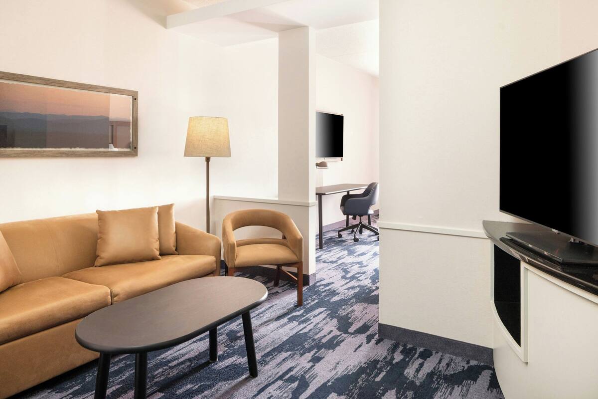 Habitación de hotel con sofá, sillón, una mesita, lámpara, televisor y escritorio con silla.