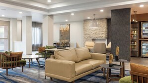 Een moderne hotel lobby met een beige bank, groene fauteuils, een eetgedeelte en een keukenruimte met een koelkast vol drankjes.