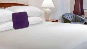 Una cama bien hecha con sábanas blancas y una almohada morada, cabecera de madera, lámpara de noche y una silla con una mesita de noche junto a la cama.
