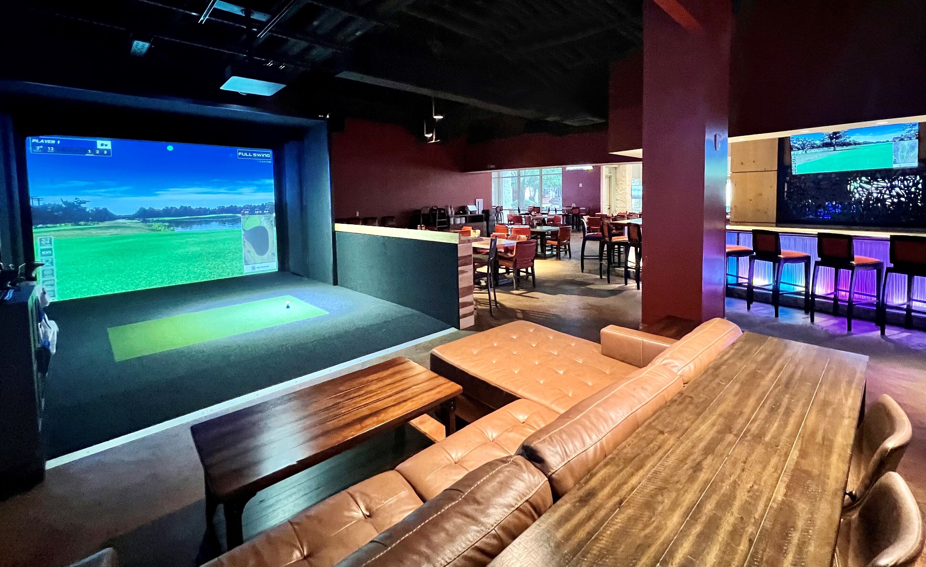 Un área de descanso moderna con una pantalla grande mostrando un partido de golf, asientos de cuero y una zona de bar con iluminación de colores.